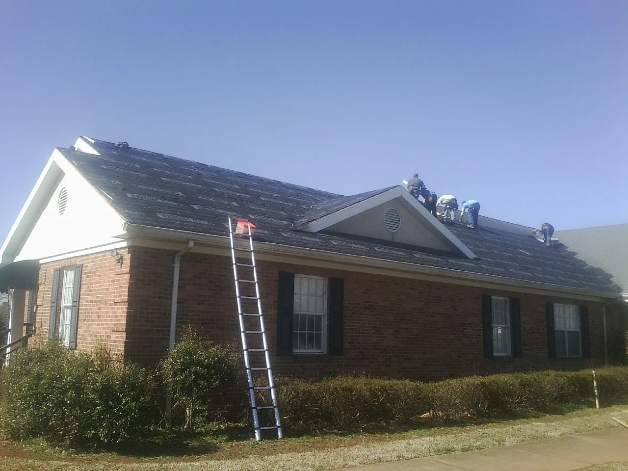 Crew tearing shingles off a brick ranch roof with a ladder set — Metal Roof Repair work by our crew in Buckland